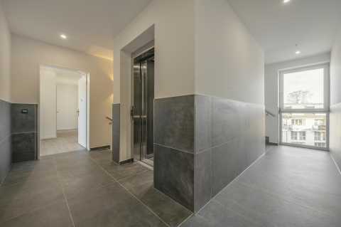 Moderner Hausflur mit Aufzug, grauen Fliesen und großem Fenster.