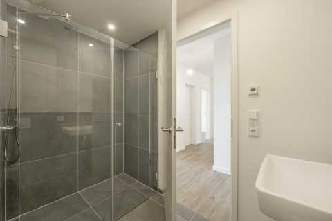 Modernes Bad mit großer Dusche, grauen Fliesen und offener Tür zum Flur.