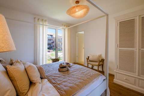 Helles Schlafzimmer mit Doppelbett, Sessel, Schrank und Fenster mit Vorhängen.