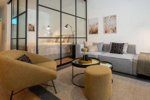 Modernes Wohnzimmer mit Glaswand, Sofa, Sessel und Blick ins Schlafzimmer.