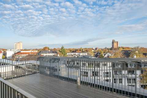 Blick von einem Balkon über eine Stadt mit Häusern, Bäumen und blauem Himmel.