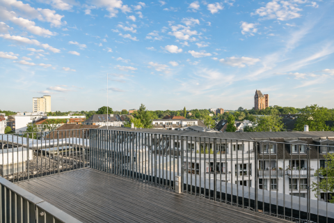 Balkon mit Metallgeländer, Blick über Häuser auf grüne Bäume und blauen Himmel.
