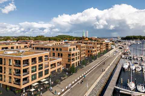 Moderne Wohnhäuser und Yachthafen an einer Promenade bei sonnigem Wetter.