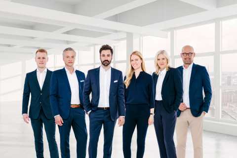 Sechs Menschen in Businesskleidung stehen in einem modernen, hellen Büro.