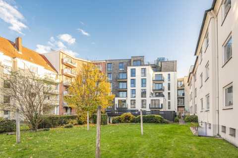 Moderner Wohnblock mit Balkonen, Wiese und Bäumen unter blauem Himmel.