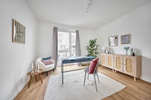 Helles Arbeitszimmer mit Schreibtisch, Stuhl, Sessel, Teppich und Kommode.