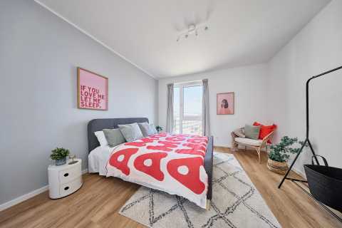 Helles Schlafzimmer mit Doppelbett, roter Decke, Stuhl und großen Fenstern.