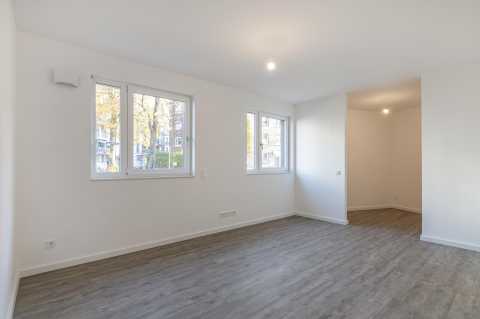 Helles, leeres Zimmer mit zwei Fenstern, weißer Wand und grauem Boden.